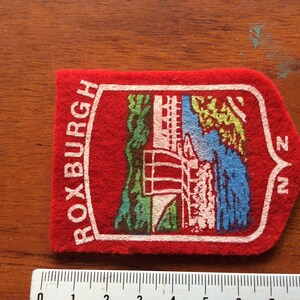 May include: Red felt patch with white text that reads "ROXBURGH NZ". The patch features a colorful illustration of a building with a bridge and trees.
