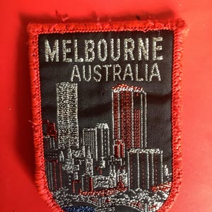 May include: A rectangular patch with a red border and the words "MELBOURNE AUSTRALIA" in silver lettering. The patch features a cityscape with silver, red, and blue details, likely a souvenir or collectible item.