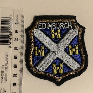 Op de afbeelding: Geborduurde patch met de tekst "EDINBURGH" boven een blauw-wit ontwerp met gele kasteeldetails. De schildvormige patch heeft een gouden rand en een zwarte achterkant. De patch is ongeveer 2,5 cm breed.