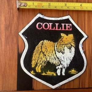 Peut inclure: Un écusson en feutre noir avec une bordure blanche en forme de bouclier. L'écusson représente un chien Collie jaune et blanc avec le mot "COLLIE" imprimé en lettres roses en haut.