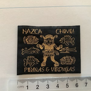 Può includere: Toppa tessuta nera con ricamo dorato raffigurante una figura stilizzata, pesci e testo. Il testo recita "NAZCA CHIMU PIRANAS & VIRONGAS". Un righello con segni in centimetri è in basso.