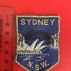 Op de afbeelding: Een blauwe geborduurde patch met het Sydney Opera House en de Harbour Bridge. De patch heeft het woord "SYDNEY" bovenaan en "N.S.W." onderaan in gouden letters. De patch is omrand met gouden stiksels.