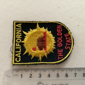 May include: A black and yellow embroidered patch with a yellow sun and a brown bear inside. The text "CALIFORNIA THE GOLDEN STATE" is embroidered in red on the patch.