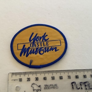 以下が含まれることがあります： 黄色い背景に青い文字で「York Castle Museum」と書かれた楕円形のパッチ。青い縁取りがあり、下に定規が置かれています。