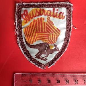 Può includere: Toppa a forma di scudo con la scritta "Australia" in giallo. La toppa presenta un canguro e un disegno a strisce rosse e gialle. Il bordo è rosso scuro con cuciture bianche.