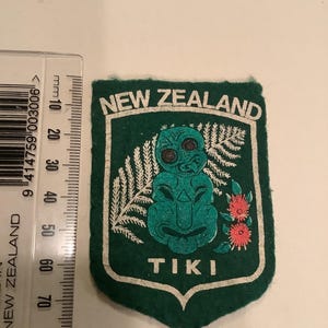 May include: A green felt patch with white trim, featuring the words "NEW ZEALAND" and "TIKI". The patch has a shield shape and includes a teal Tiki figure, white fern, and red flowers.