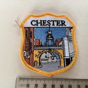 Op de afbeelding: Geborduurde patch in schildvorm met het woord "CHESTER" bovenaan. De patch toont een kleurrijke illustratie van gebouwen, een brug en een klokkentoren. De rand is geel.