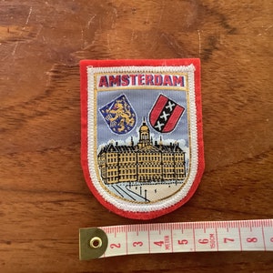 May include: Red, white and blue embroidered patch with the word "AMSTERDAM" in white lettering. The patch features a building with two coats of arms, one with a lion and the other with an "XXX" symbol.