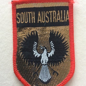 May include: A shield-shaped patch with a red border. The patch features the text "SOUTH AUSTRALIA" above a black eagle with white and blue details, set against a gold and black background.