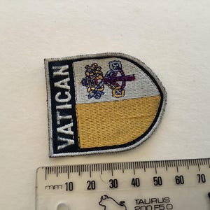 Könnte beinhalten: Gestickter Aufnäher mit dem Wort "VATICAN" in Schwarz auf weißem und gelbem Hintergrund. Der Aufnäher zeigt ein Wappen mit religiösen Symbolen. Ein Lineal ist darunter platziert und zeigt die Maße in Millimetern an.