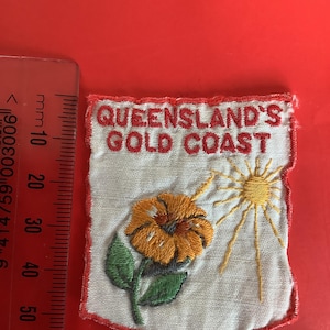 May include: Embroidered patch with the text "QUEENSLAND'S GOLD COAST". The patch features a yellow flower, green leaves, and a yellow sun. The patch has a red border and is on a white background.