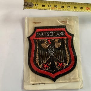 Puede incluir: Parche bordado con la palabra "DEUTSCHLAND" sobre un emblema de águila negra sobre un escudo. El parche tiene una base negra con detalles en rojo y dorado. El parche mide aproximadamente 7.6 cm de ancho.