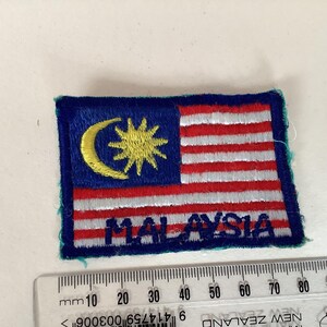 May include: Embroidered patch featuring the flag of Malaysia. The flag design includes a blue field with a yellow crescent and star, and red and white stripes. The word "MALAYSIA" is embroidered below the flag.