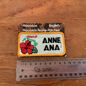 Può includere: Una toppa bianca ricamata con un bordo giallo. La toppa presenta un fiore di ibisco rosso, foglie verdi e il testo "Hawaii" in corsivo. Il testo "ANNE ANA" è ricamato in nero sotto il fiore. La toppa è confezionata su una carta con il testo "Hawaiian Name Patches" in nero.