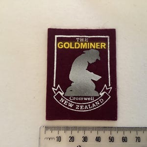 May include: A maroon patch with the words "THE GOLDMINER" in gold lettering. Below is a silver silhouette of a person, and the words "Cromwell NEW ZEALAND" in white. A ruler is at the bottom.