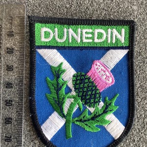 May include: Embroidered patch featuring the word "DUNEDIN" in white on a green background. The patch has a blue field with a white cross, a green thistle, and a pink flower. The patch is bordered in black.