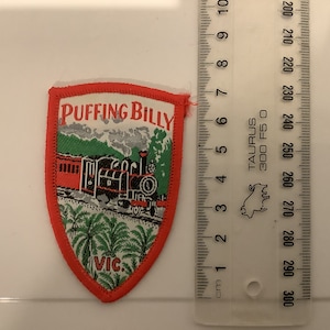 May include: Shield-shaped patch with a red border featuring a train illustration. The train is black and red, with the words "PUFFING BILLY" at the top and "VIC" at the bottom. The background includes green hills and palm trees.