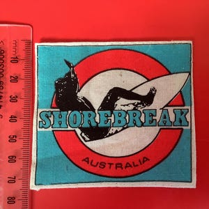 Puede incluir: Un parche rectangular con un gráfico de surfista. El parche presenta una silueta negra de un surfista sobre una tabla de surf blanca, sobre un fondo circular rojo y blanco. Se muestra el texto "SHOREBREAK AUSTRALIA".