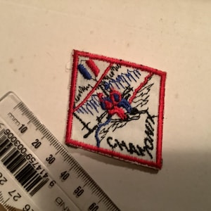 May include: Square embroidered patch with a red border and a white centre. The design features a figure in red and blue, with black and blue lines. The word "CHAF" is visible. A ruler is in the image.