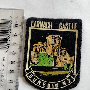 May include: A black patch with a gold and green illustration of Larnach Castle, with the text "Larnach Castle Dunedin N.Z." The patch is approximately 7.6 cm wide and 7.6 cm tall.