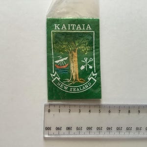 Könnte beinhalten: Ein grüner Aufnäher mit den Worten "Kaitaia" und "Neuseeland" in weißen Buchstaben. Der Aufnäher zeigt einen stilisierten Baum mit einem Boot und einer Schaufel im Hintergrund.