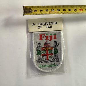 May include: A souvenir patch from Fiji, featuring the country's coat of arms and the word "Fantastic." The patch is in a clear plastic sleeve, with the text "A Souvenir of Fiji" at the top. A measuring tape is in the background.