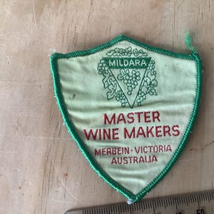 May include: A shield-shaped patch with green edging and text. The patch reads "MILDARA" above a grape design, and "MASTER WINE MAKERS" below. The text "MERBEIN - VICTORIA AUSTRALIA" is also present. The patch is on a wooden surface.