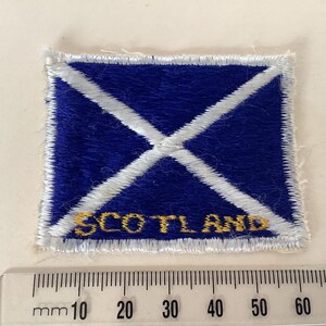 Op de afbeelding: Een rechthoekige patch met het Schotse vlagontwerp. De patch heeft een blauwe achtergrond met een witte diagonale kruis. Het woord "SCOTLAND" is in goud geborduurd. De patch is ongeveer 5 cm breed.