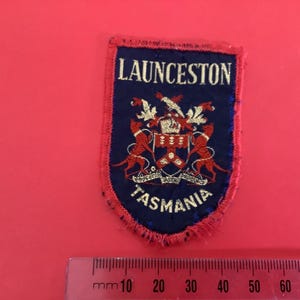 May include: A blue and red embroidered patch with the text "LAUNCESTON TASMANIA". The patch features a crest with two red lions, a crown, and other heraldic elements. The patch is approximately 3 inches wide.