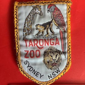 May include: Embroidered patch featuring a giraffe, monkey, and parrot, with a lion's head at the bottom. The patch reads "TARONGA ZOO SYDNEY N.S.W." in red and black lettering, framed by a gold border.