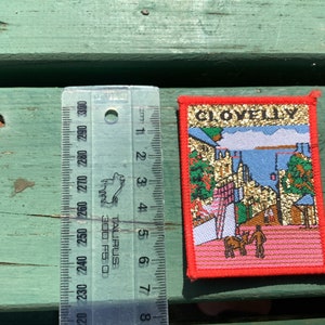 May include: A red, white, and blue embroidered patch with the word "CLOVELLY" in gold glitter lettering. The patch depicts a colorful scene of a village with buildings, a harbor, and a horse-drawn carriage.