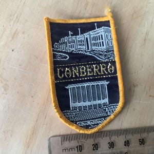 May include: A navy blue and gold embroidered patch with the word "CONBERRO" in gold lettering. The patch features a detailed illustration of a building and a landscape design. The patch has a yellow border.