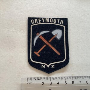 May include: Black and white patch with the text "GREYMOUTH NZ" in white. The patch features a stylised image of a pickaxe and shovel crossed over each other.