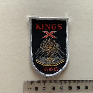 Könnte beinhalten: Gestickter Aufnäher mit schwarzem Hintergrund, mit den roten Worten "KINGS X" und einem Gold- und Silberdesign. Das Wort "SYDNEY" steht unten. Der Aufnäher hat einen weißen Rand.