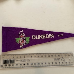 Puede incluir: Bandera de fieltro morado con la palabra "DUNEDIN" en blanco y "N-Z". Presenta una imagen bordada de una persona tocando la gaita. Una regla en cm está debajo.