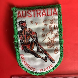 May include: A patch with a green border and the word "AUSTRALIA" in red. The patch features an illustration of an Aboriginal didgeridoo player in red, black, and brown, with the words "DIDJERIDOO PLAYER" below.