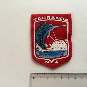 May include: A red patch with a shield design featuring the word "TAURANGA" at the top. The shield contains a blue marlin leaping from blue waves, with a white boat in the background. The letters "NZ" are at the bottom.
