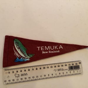 Może przedstawiać: Bordowa chorągiewka z grafiką skaczącej ryby i napisem "TEMUKA New Zealand". Obok chorągiewki znajduje się linijka. Chorągiewka ma około 18 cm długości.