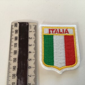 May include: An embroidered iron-on patch featuring the Italian flag with the word "ITALIA" in red lettering. The patch is approximately 6 cm wide.