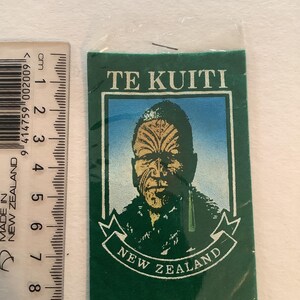 May include: A green rectangular souvenir featuring a portrait of a person with traditional facial tattoos. The text "TE KUITI" is at the top, and "NEW ZEALAND" is at the bottom. A ruler is next to the souvenir, showing measurements in centimeters.
