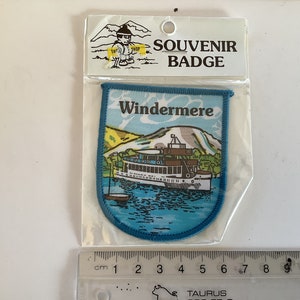 Op de afbeelding: Blauw en wit souvenirbadge met de tekst "SOUVENIR BADGE" en "Windermere". De badge heeft een cartoonillustratie van een boot op een meer met bergen op de achtergrond.