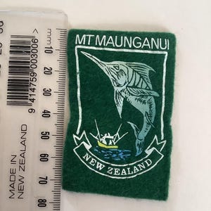 May include: Green felt patch featuring a marlin illustration and the text "MT MAUNGANUI" and "NEW ZEALAND" in white. A ruler is placed next to the patch. The patch is approximately 7 cm in length.