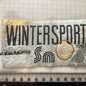 Può includere: Una toppa di tessuto bianco con testo nero che recita "WINTERSPORT" e una grafica nera e gialla di una "S" stilizzata.