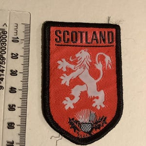 Pode incluir: Um patch vermelho e preto com a palavra "SCOTLAND" no topo. O patch apresenta um leão rampante branco e um emblema de cardo. O patch tem formato de escudo e tem uma borda preta.