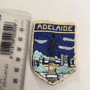May include: A blue embroidered patch with the word "ADELAIDE" at the top. The patch features a cityscape with a statue and clouds. The patch is approximately 2 inches wide and 2.5 inches tall.