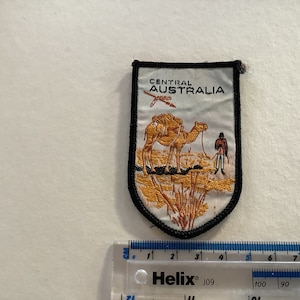 May include: A white embroidered patch with a camel and a person walking in the desert. The patch has black stitching around the edges and says "Central Australia" in black text.