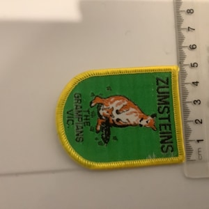 Op de afbeelding: Een rechthoekige patch met een gele rand en een groene achtergrond. De patch toont een bruin-witte dierenillustratie en de tekst "ZUMSTEINS" en "THE GRAMPIANS VIC." De patch is ongeveer 5 cm lang.