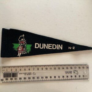 Puede incluir: Bandera de fieltro negro con la palabra "DUNEDIN" en blanco, "N.Z." y una ilustración de una persona con vestimenta escocesa. Una regla está colocada debajo. La bandera mide unos 18 cm de largo.