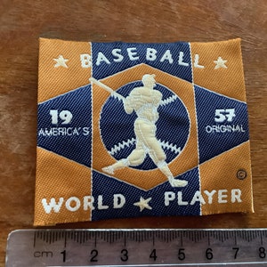 Puede incluir: Un parche de béisbol vintage con una silueta blanca de un jugador de béisbol sobre un fondo azul y naranja. El parche está bordado con el texto "BASEBALL", "19 AMERICA'S", "57 ORIGINAL", "WORLD" y "PLAYER" en blanco.