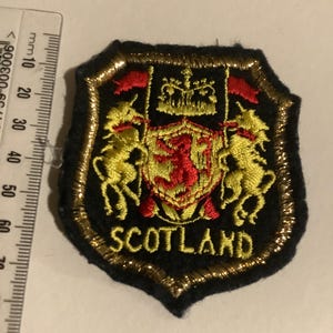 May include: Embroidered patch featuring the Scottish coat of arms. The patch is shield-shaped with a black background and gold trim. The design includes a red lion rampant, gold unicorns, and the word "SCOTLAND".